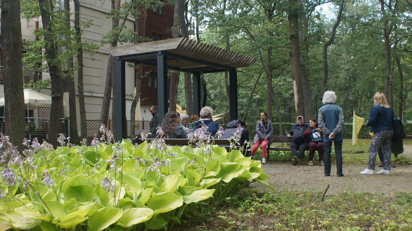 Na zdjęciu widać park oraz ludzi stojących i siedzących ma ławeczkach.