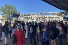 Uczestnicy wycieczki na zbiórce, słuchający wytycznych przewodników. W tle stadion Łódzkiego Klubu Sportowego