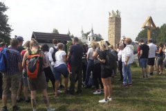 Grupa osób stoi na trawie przed turystyczną tablicą informacyjną w tle dwie wieże kościoła, pomnik z koroną na szczycie, drewniany krzyż i autokar