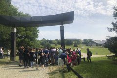 Grupa osób stoi przed bramą do ośrodka rekreacyjnego, w tle drzewa, trawa  i domki noclegowe