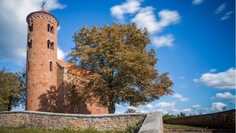 Ceglany kościół usytuowany po lewej stronie zdjęcia, przed nim kamienny murek oraz po prawej stronie zdjęcia kamienne schody. Po prawej stronie kościoła, w centralnej części zdjęcia znajduje się duże drzewo liściaste, w tle błękitne niebo oraz chmury.