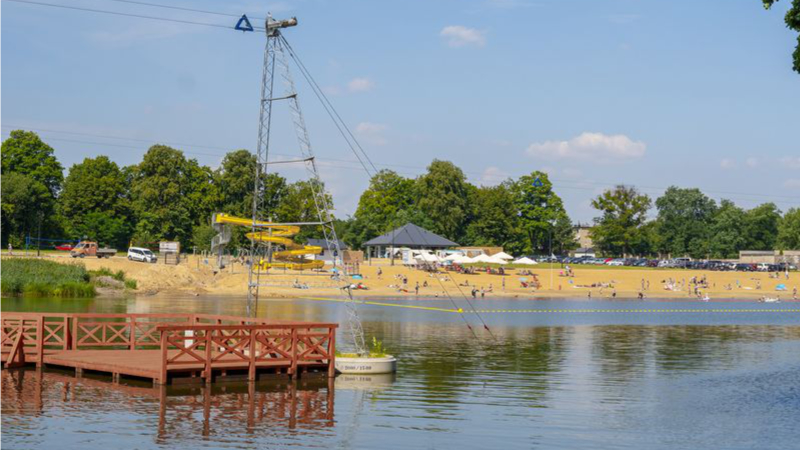 Zbiornik wodny usytuowany na pierwszym planie, w lewej części zdjęcia znajduje się drewniane molo w kolorze brązowym. Za molo znajduje się słup z linami do wake parku. w dalszym panie widać plaże z ludźmi oraz zielone drzewa.