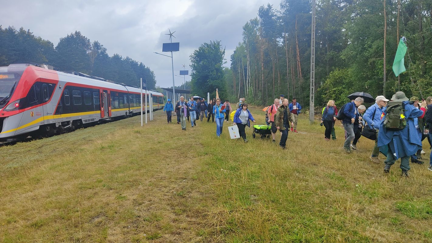 Po lewej stronie stoi pociąg, w centrum i po prawej stronie zdjęcia widać ludzi wysiadających z pociągu. Stacja w otoczeniu zieleni i lasu.
