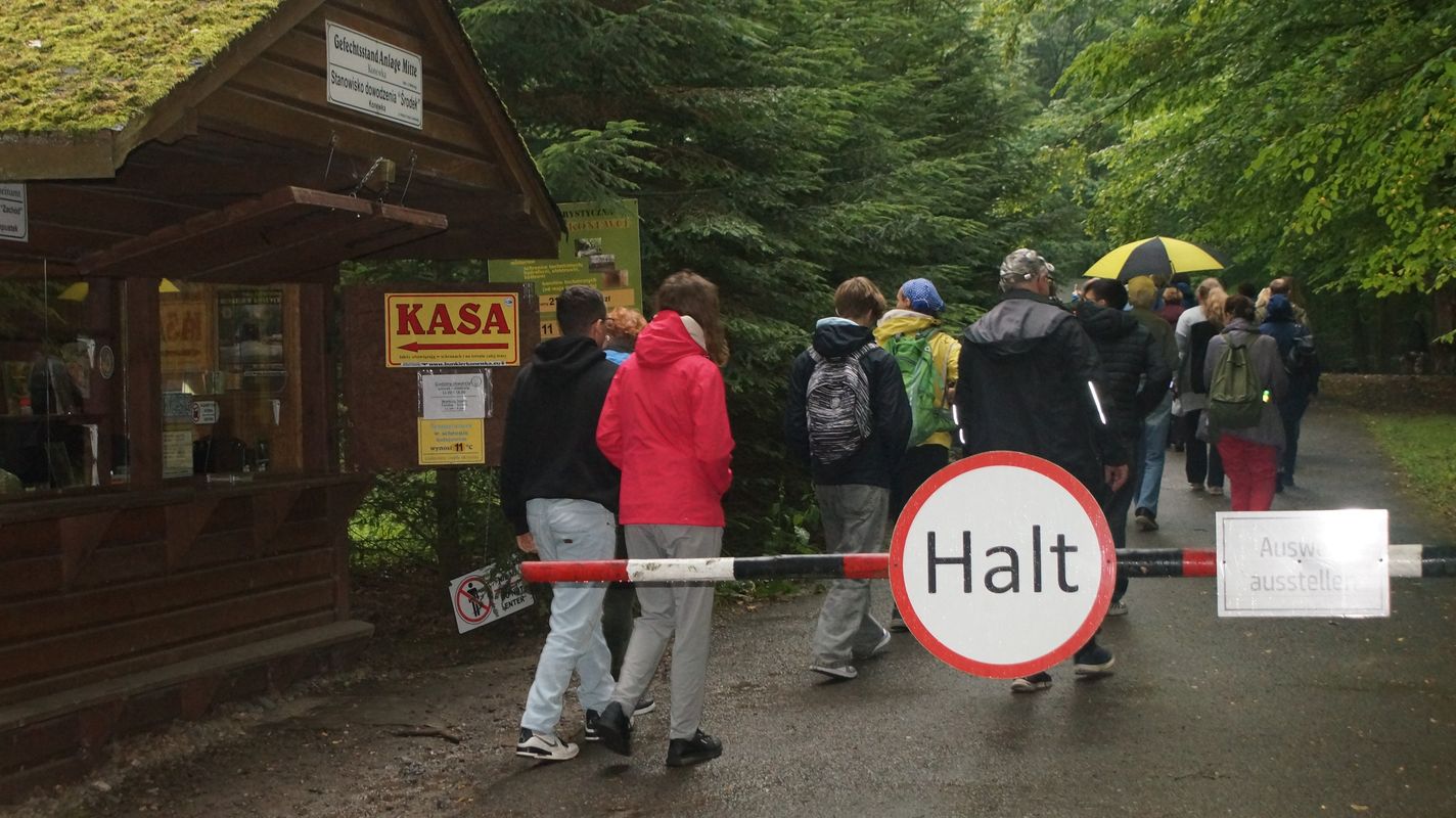 Po lewej stronie widać kiosk z biletami do muzeum. W centralnej części zdjęcia widać szlaban i drogę, po której idą ludzie.