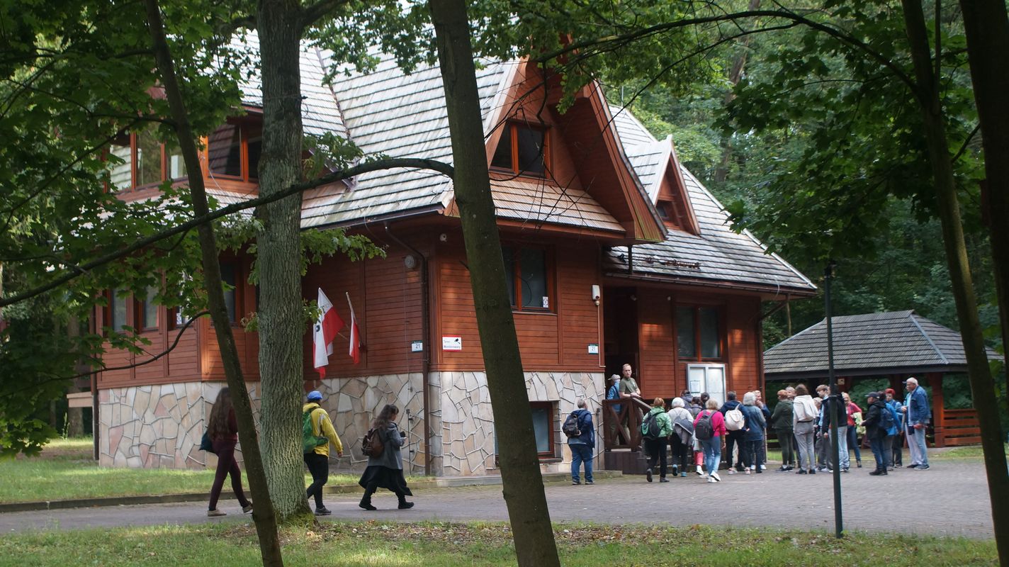 W centralnej części zdjęcia widać drewniany budynek w kolorze brązowym w otoczeniu drzew. Przed budynkiem widać stojących ludzi.