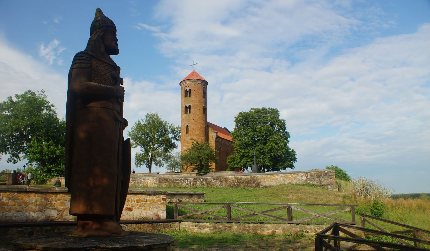 Na pierwszym planie po lewej stronie zdjęcia widać drewnianą rzeźbę, natomiast w oddali widać romański kościółek.
