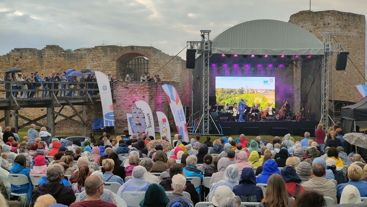 Na zdjęciu widać ludzi siedzących na krzesłach oraz stojących na mostku. W oddali widać scenę ustawioną na tle zamku. Na scenie odbywa się koncert.