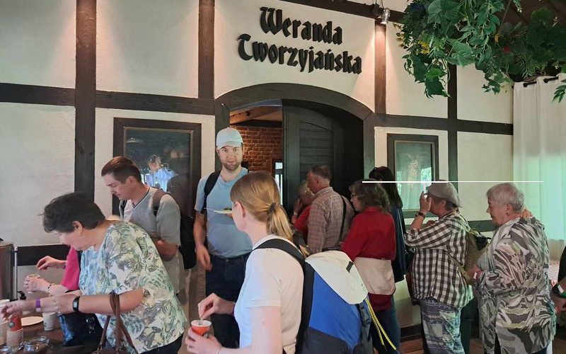 Grupa ludzi częstuje się herbatą i stoi w kolejce. W tle na górze widać napis „Weranda Tworzyjańska”