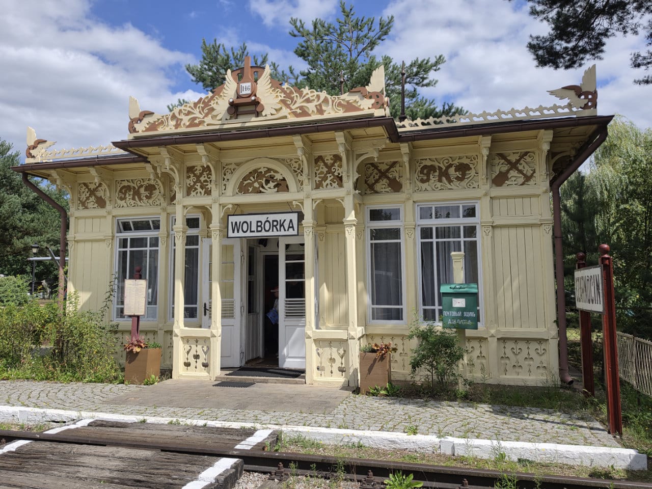 Budynek z ornamentami i tabliczką z napisem Wolbórka. W tle znajdują się zielone drzewa i zachmurzone niebo.